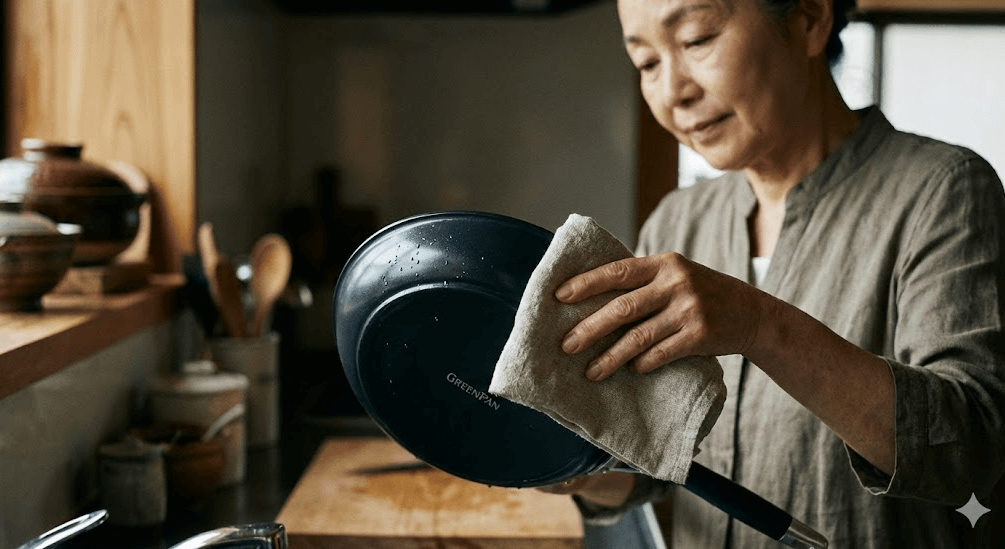 洗った後のグリーンパンを丁寧に布巾で拭き上げている年配の日本人女性。道具を大切に扱う丁寧な暮らしのワンシーン。