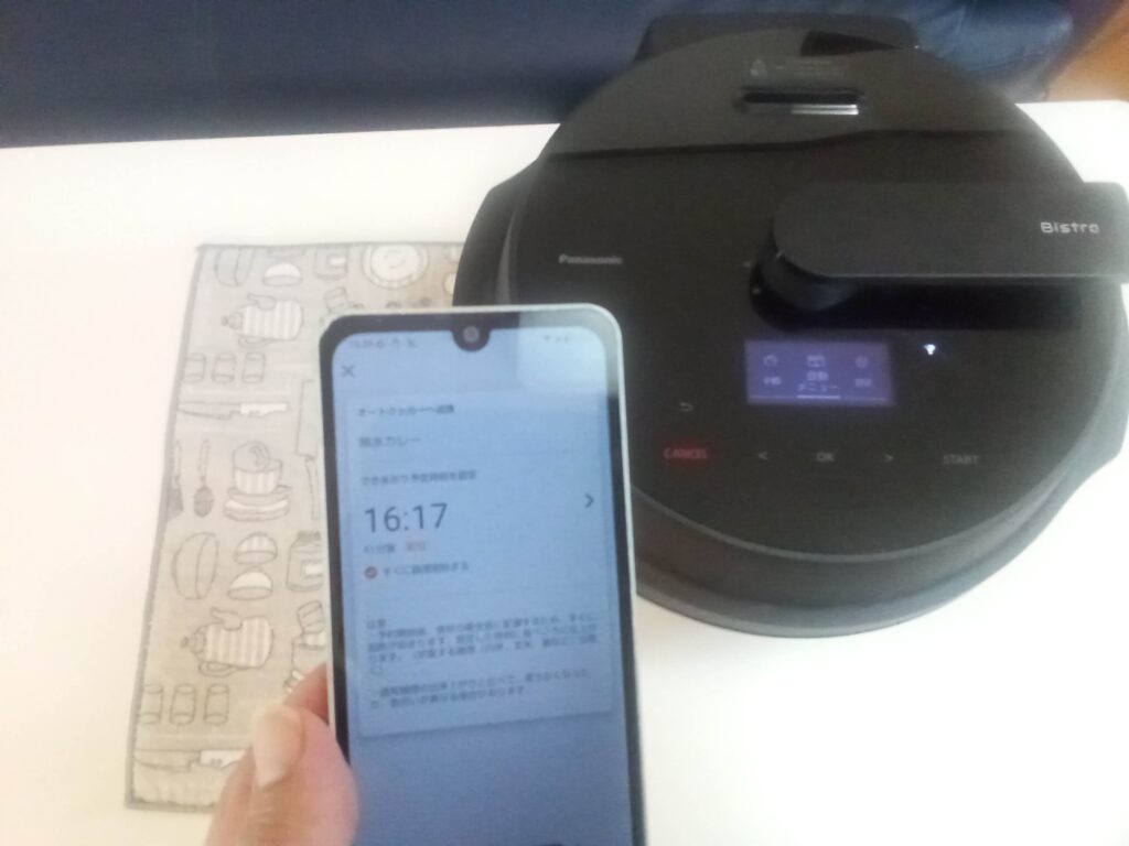 オートクッカービストロの前で、キッチンポケットアプリのできあがり予定時刻を確認するスマートフォン